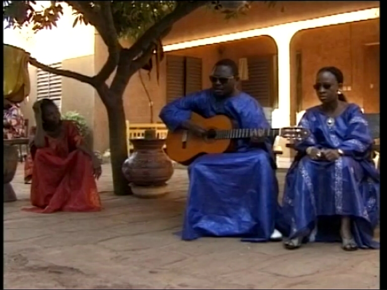 Amadou et Mariam jouant de la guitare et chantant devant une maison